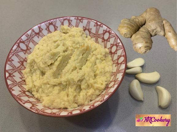 A small ceramic jar of homemade ginger garlic paste on a wooden cutting board, surrounded by fresh ginger root and garlic cloves.