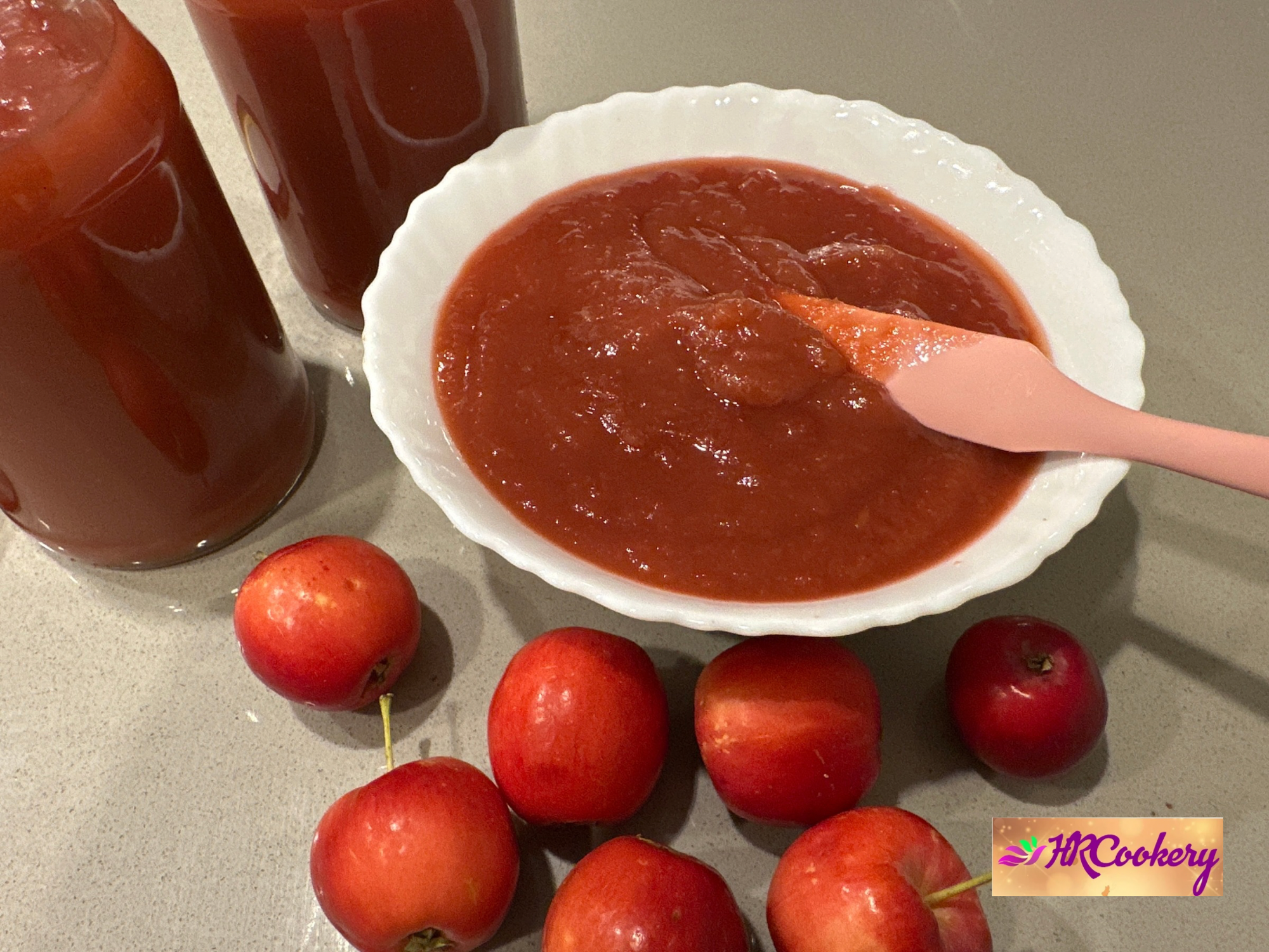 A jar of homemade apple jam with a spoon, surrounded by fresh apples and slices of toast on a rustic wooden table.